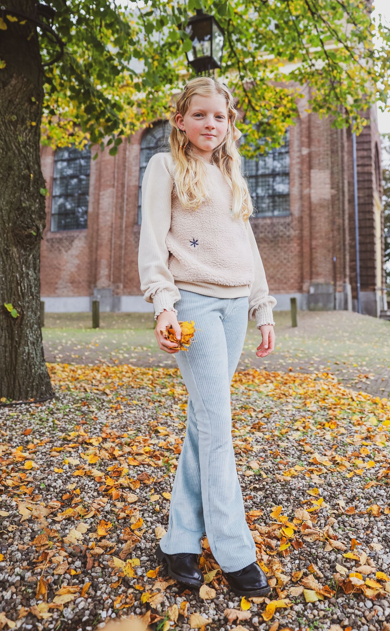 A person in a cream sweater and flared jeans poses with fall leaves outside a brick church.