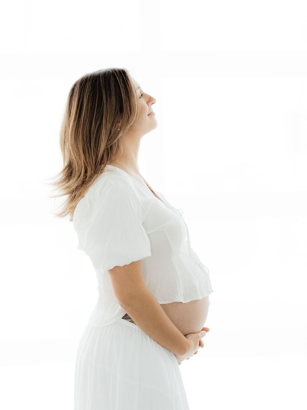 Pregnant woman in white dress holding baby bump, side profile view against white background.