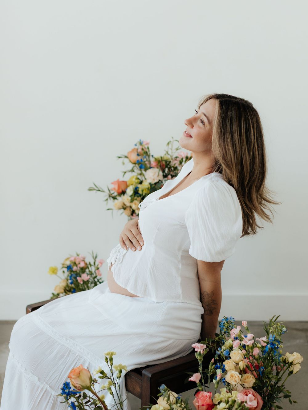 Pregnant woman in white dress sitting among colorful spring flowers, cradling her baby bump in a serene studio setting.
