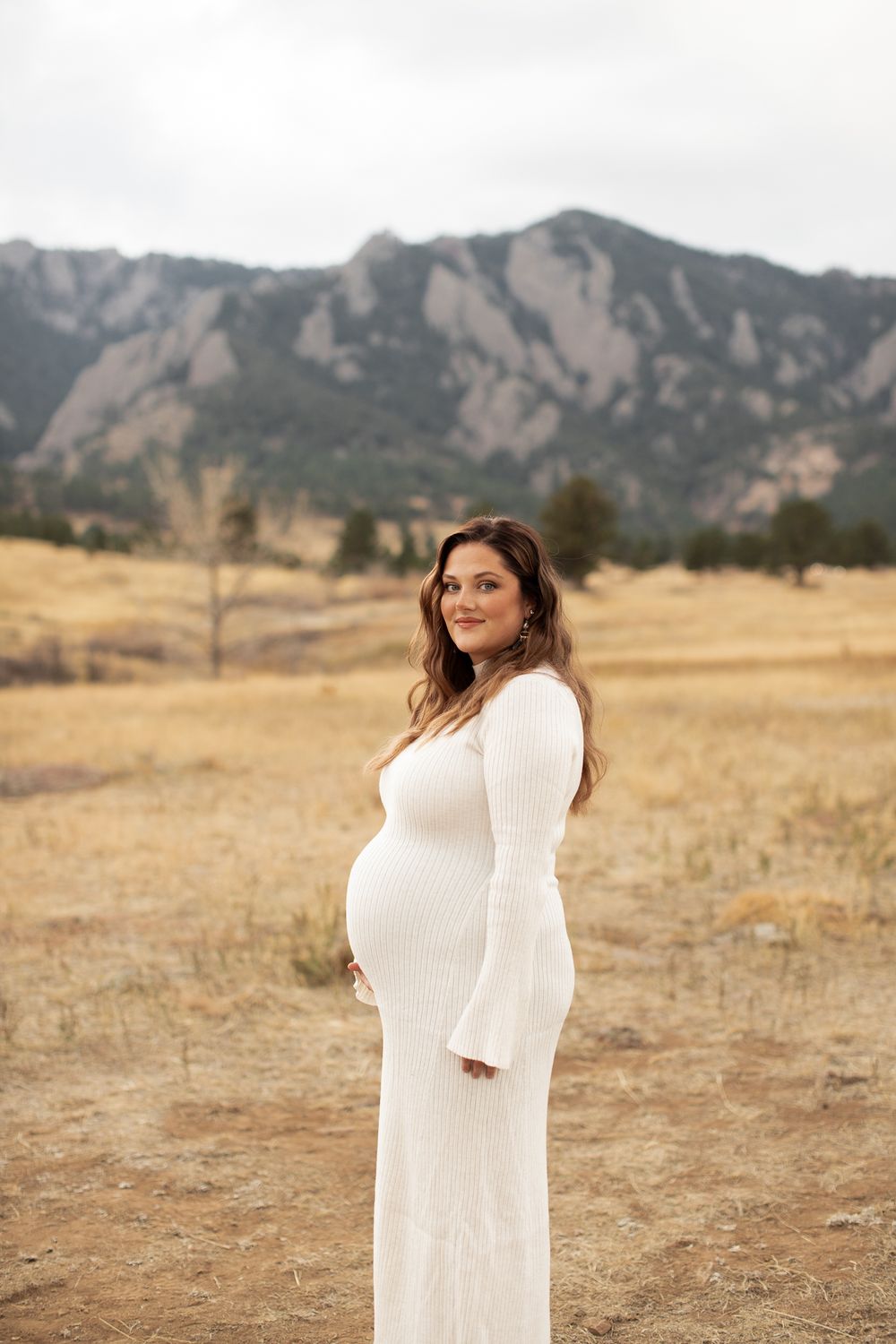 A white maternity gown adorns an expectant mother posing in a field with dramatic Flatiron mountains as backdrop in Boulder.