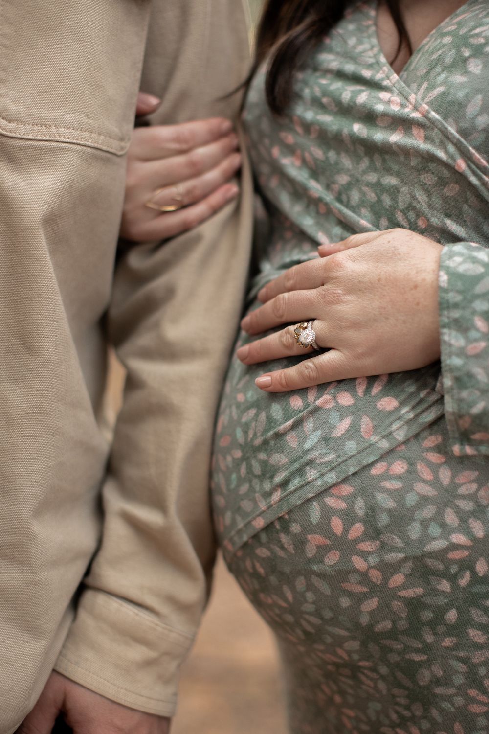 Close-up of hands showing engagement ring against sage green floral dress and tan clothing in maternity photo.