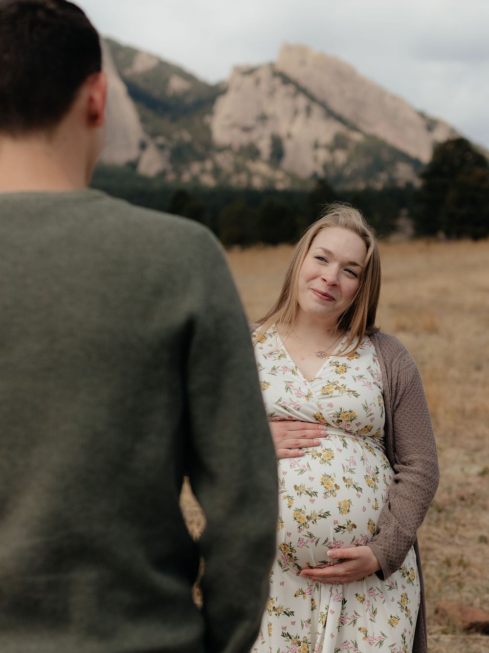 Pregnant woman in floral dress holds baby bump in mountain field during maternity photo session in Boulder, Colorado.