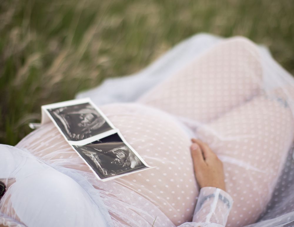 Ultrasound photos rest on a white polka dot dress against grass in a maternity photo composition.