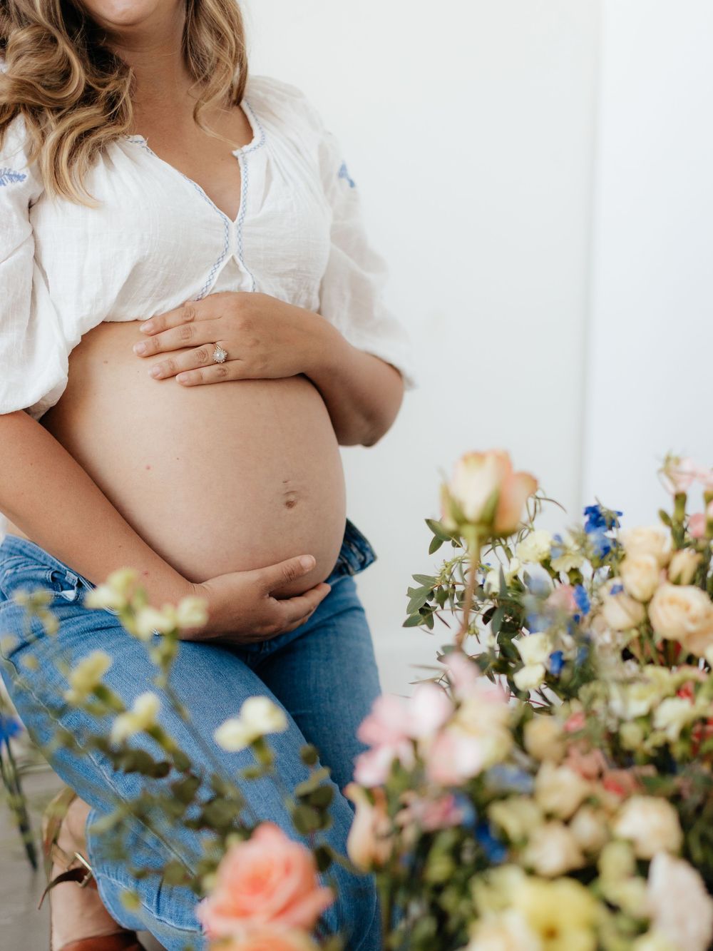 Pregnant woman in white crop top and jeans cradling baby bump surrounded by colorful spring flowers.