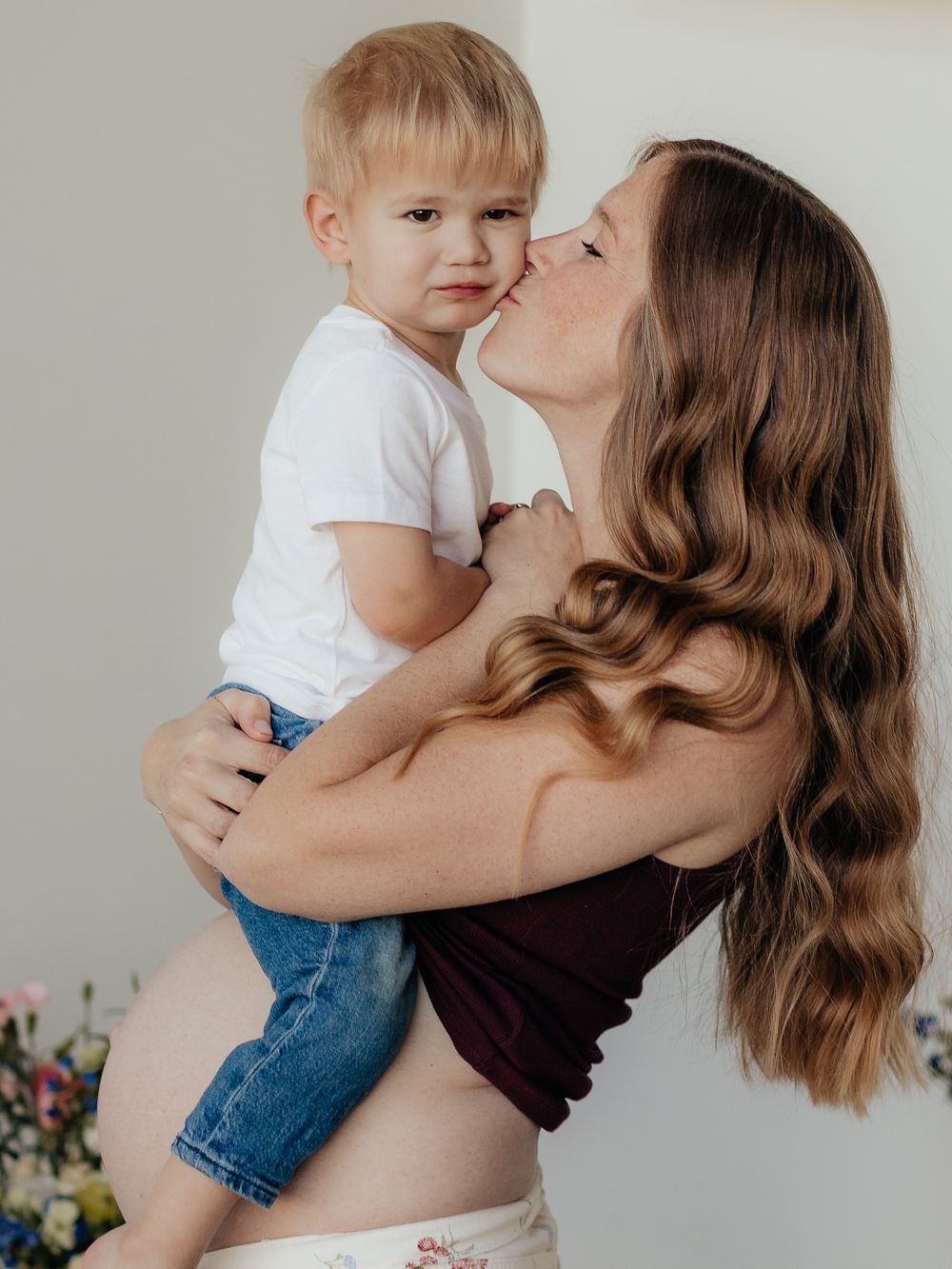 Pregnant woman with long wavy hair holding and kissing a toddler boy in white shirt and jeans.