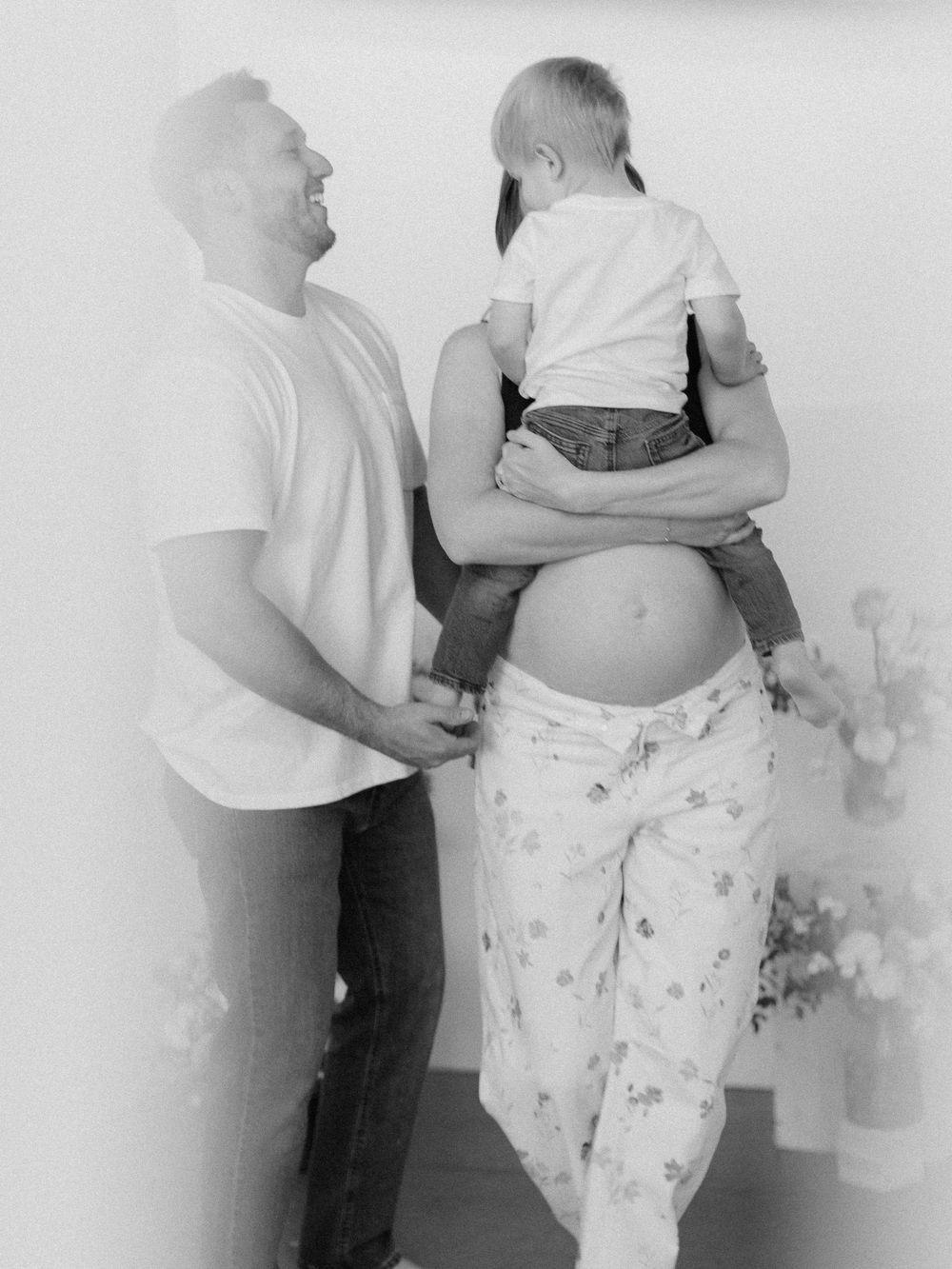 Black and white photo of a pregnant woman holding a toddler while a man stands beside her smiling.