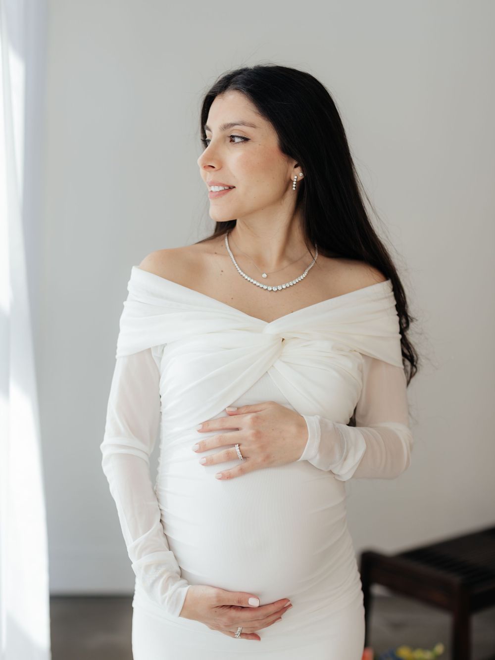 Pregnant woman in white off-shoulder gown with pearl necklace cradling baby bump, gazing softly to the side.