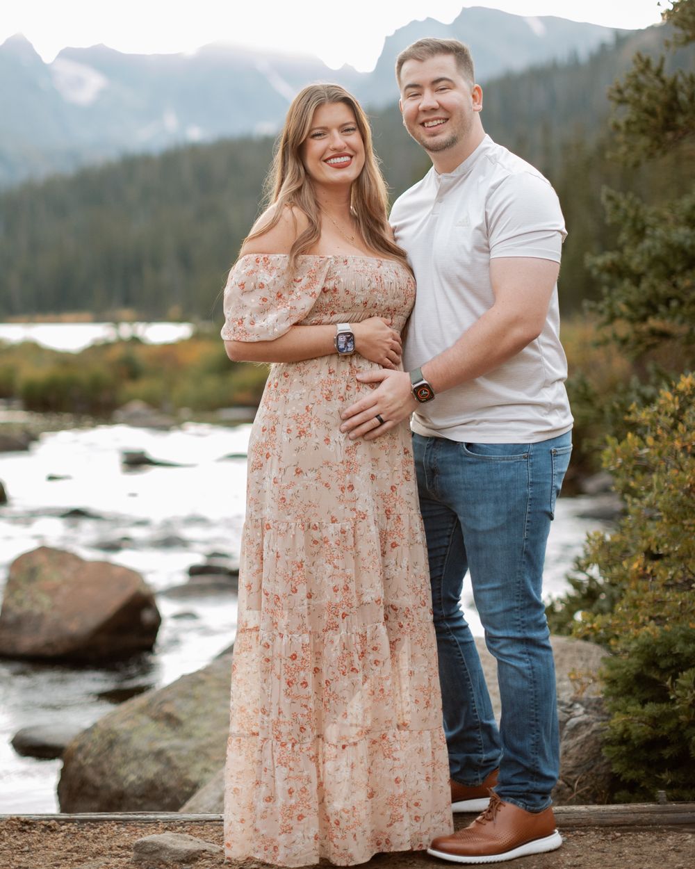 Couple poses together in front of a mountain stream wearing a floral maxi dress and white t-shirt with jeans.