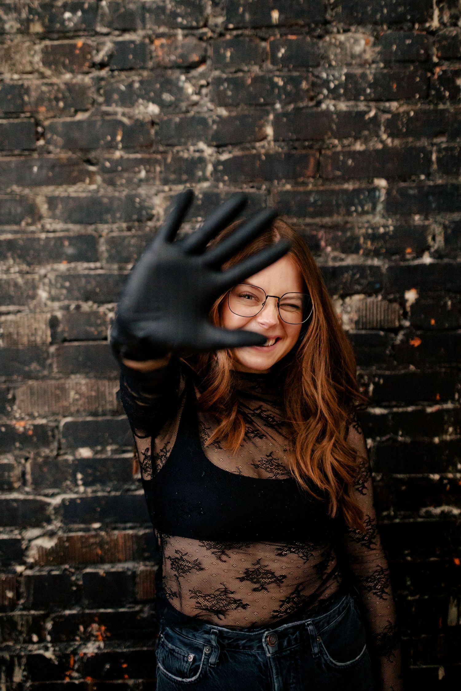 A joyful moment captured against dark brick wall as figure in lace top and denim holds up hand to camera.