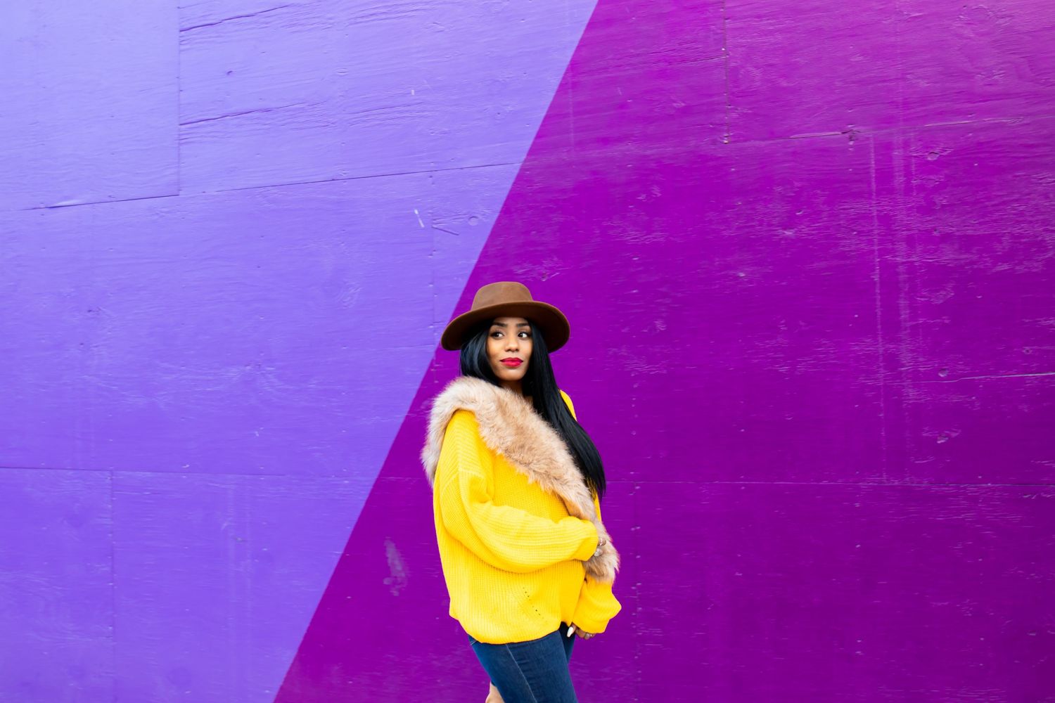 A person in a yellow sweater and black hat stands against a vibrant purple wall with diagonal color blocking.