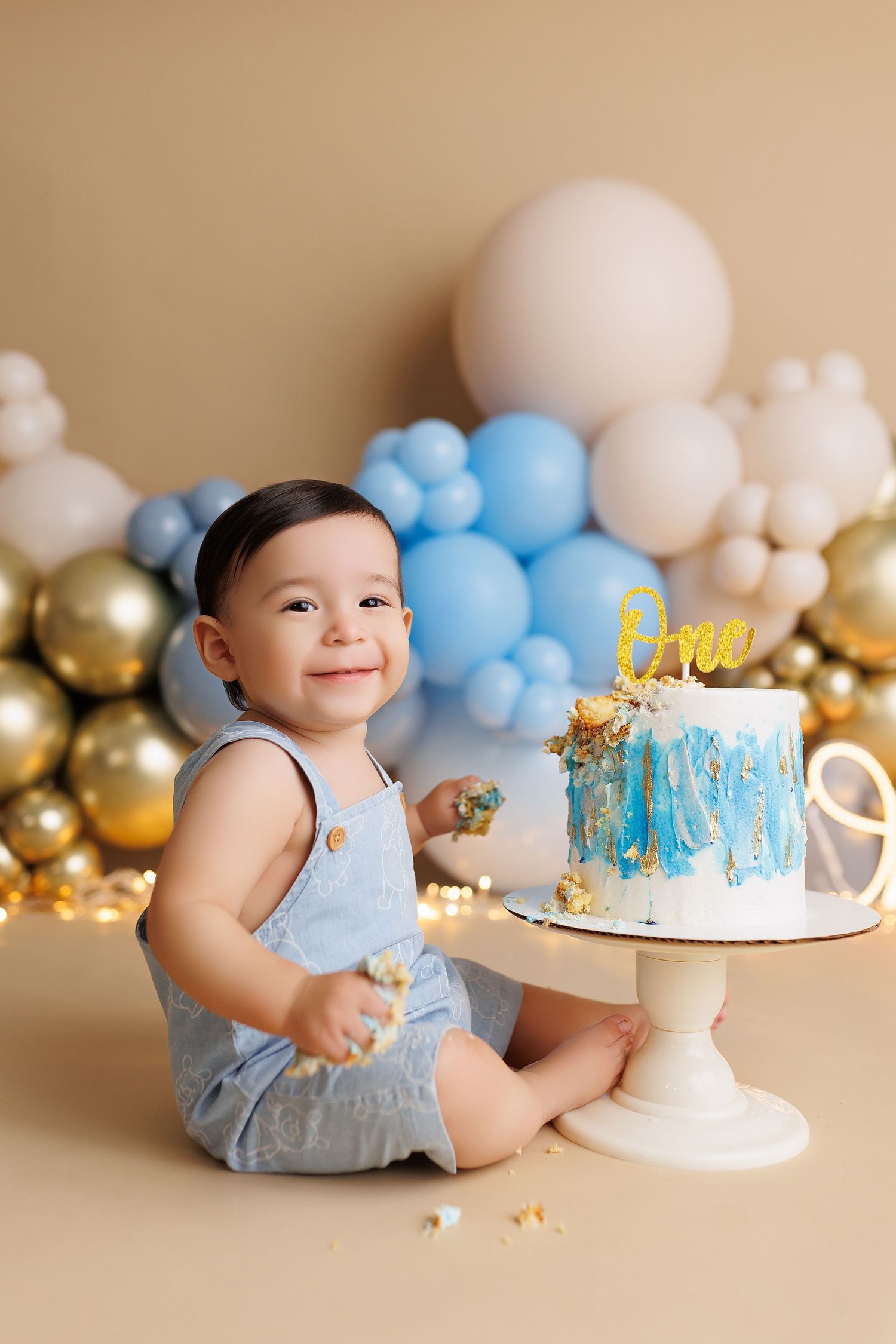 Baby boy in blue outfit enjoying first birthday cake smash photoshoot with balloon decorations.