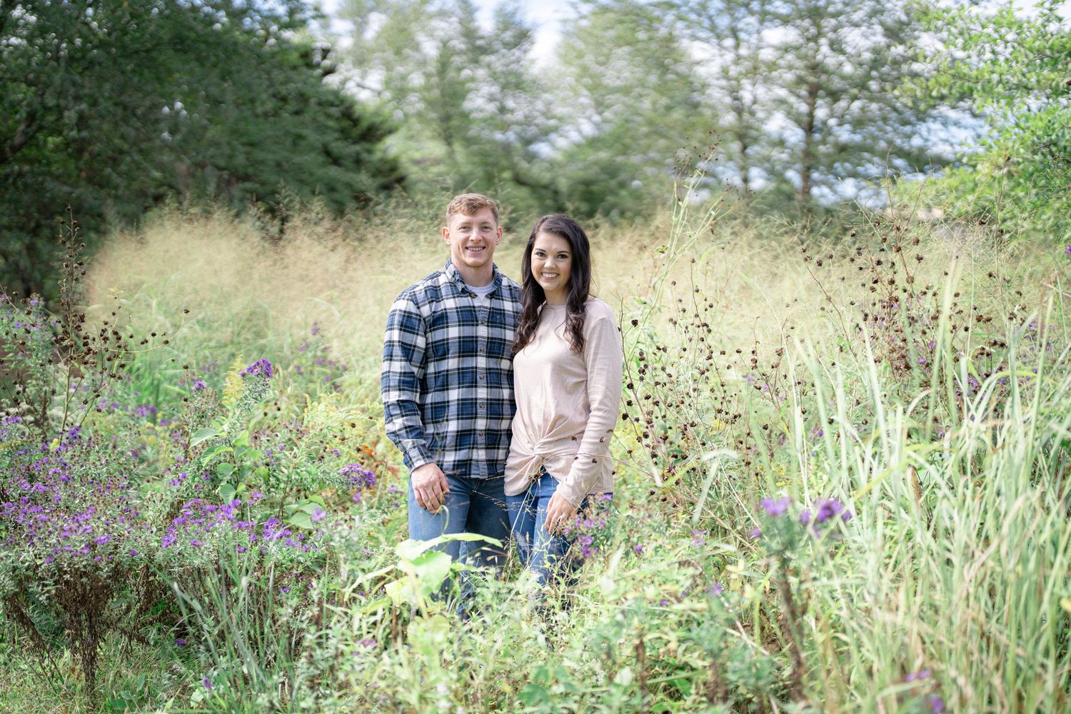 Two people stand together in a meadow with tall grass and purple wildflowers.