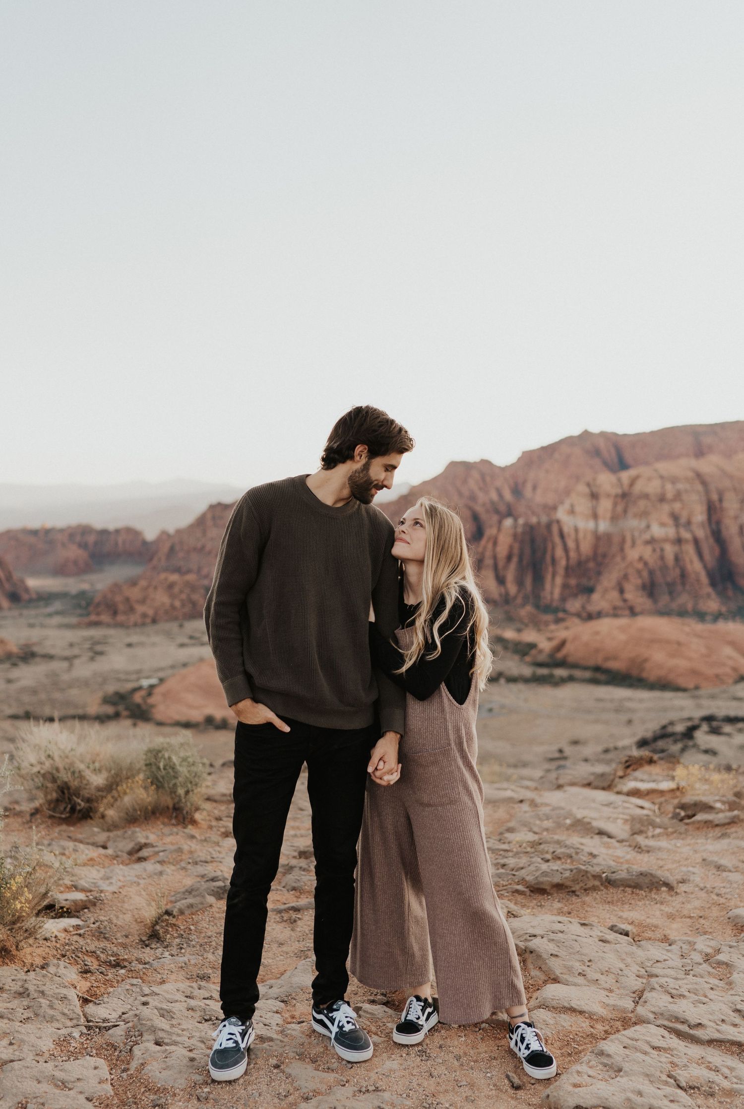 A couple in casual sweaters stands together on red rocks overlooking a desert canyon landscape.