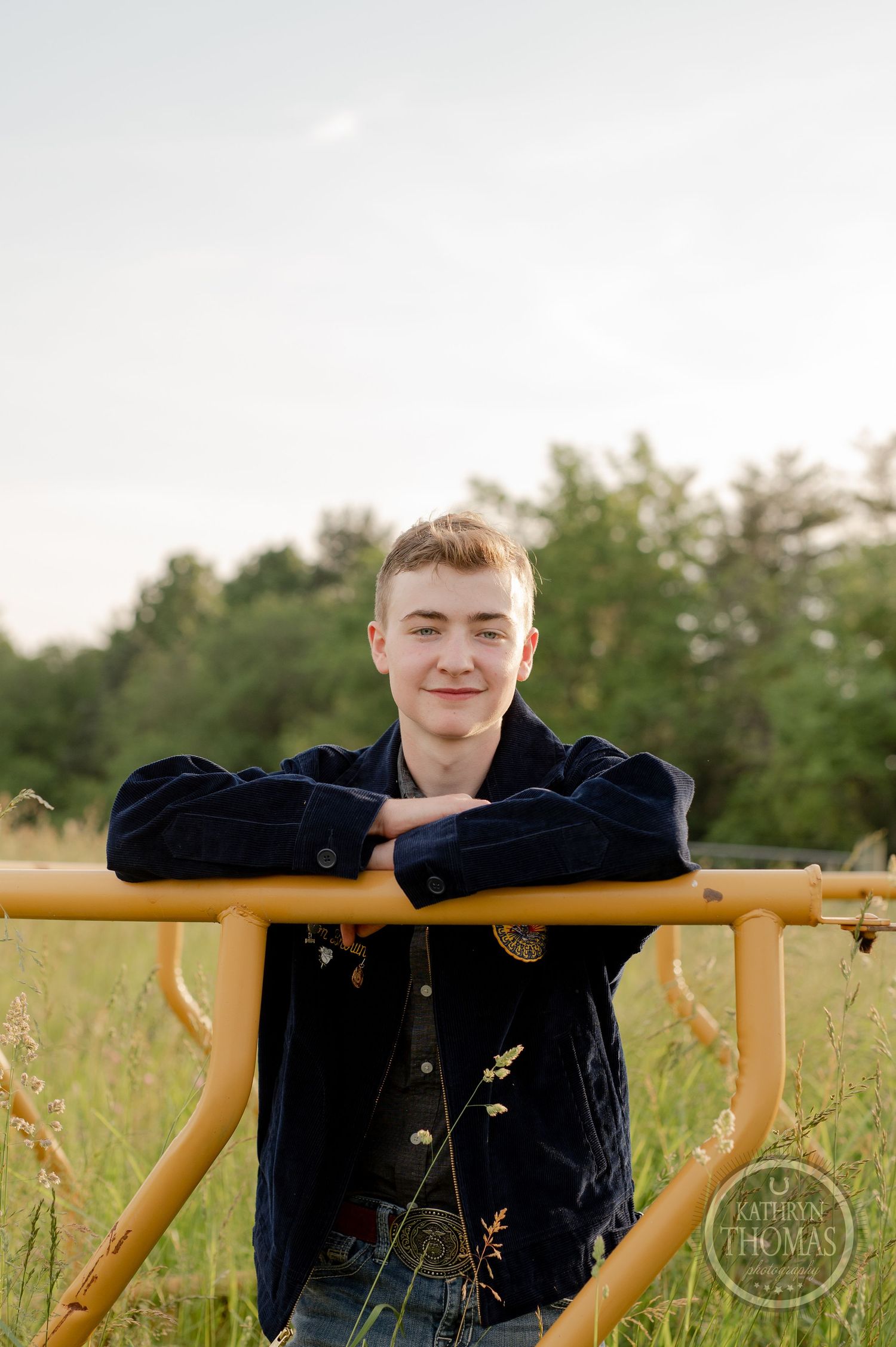 FFA member in navy jacket leans against yellow railing in grassy field during outdoor portrait session.