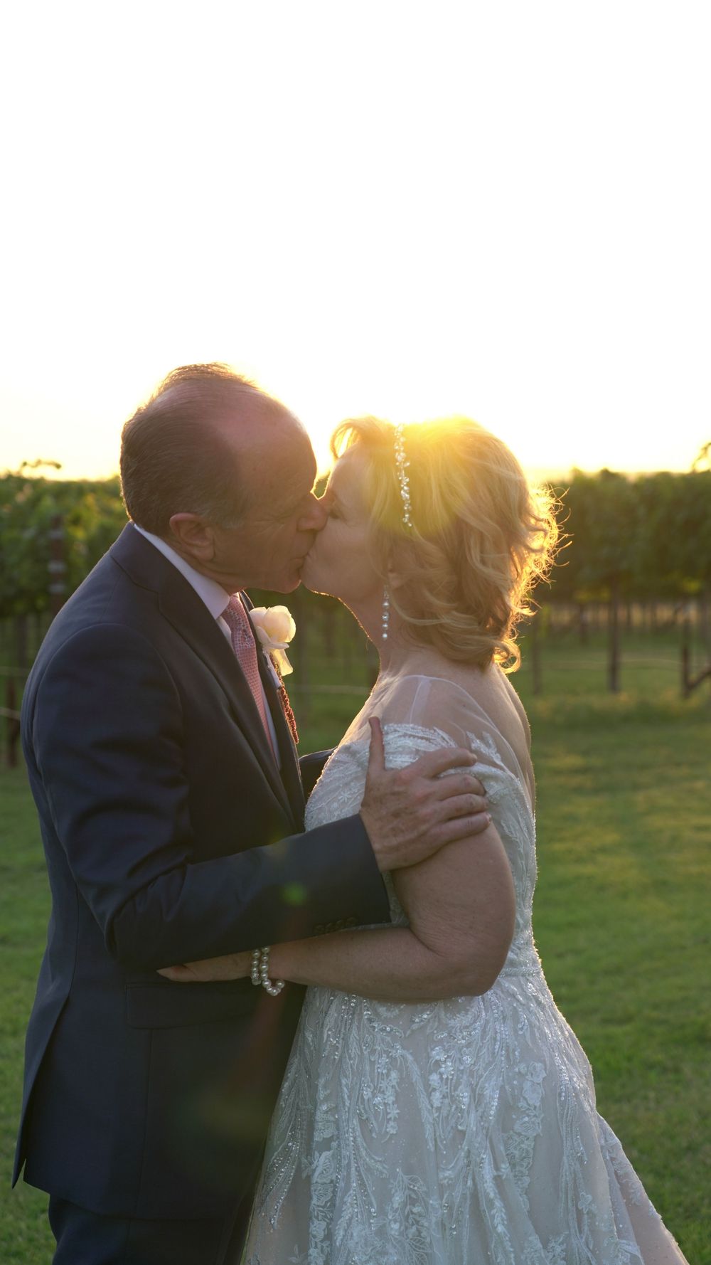 Newlywed couple sharing a romantic kiss at sunset in a vineyard while wearing formal wedding attire.