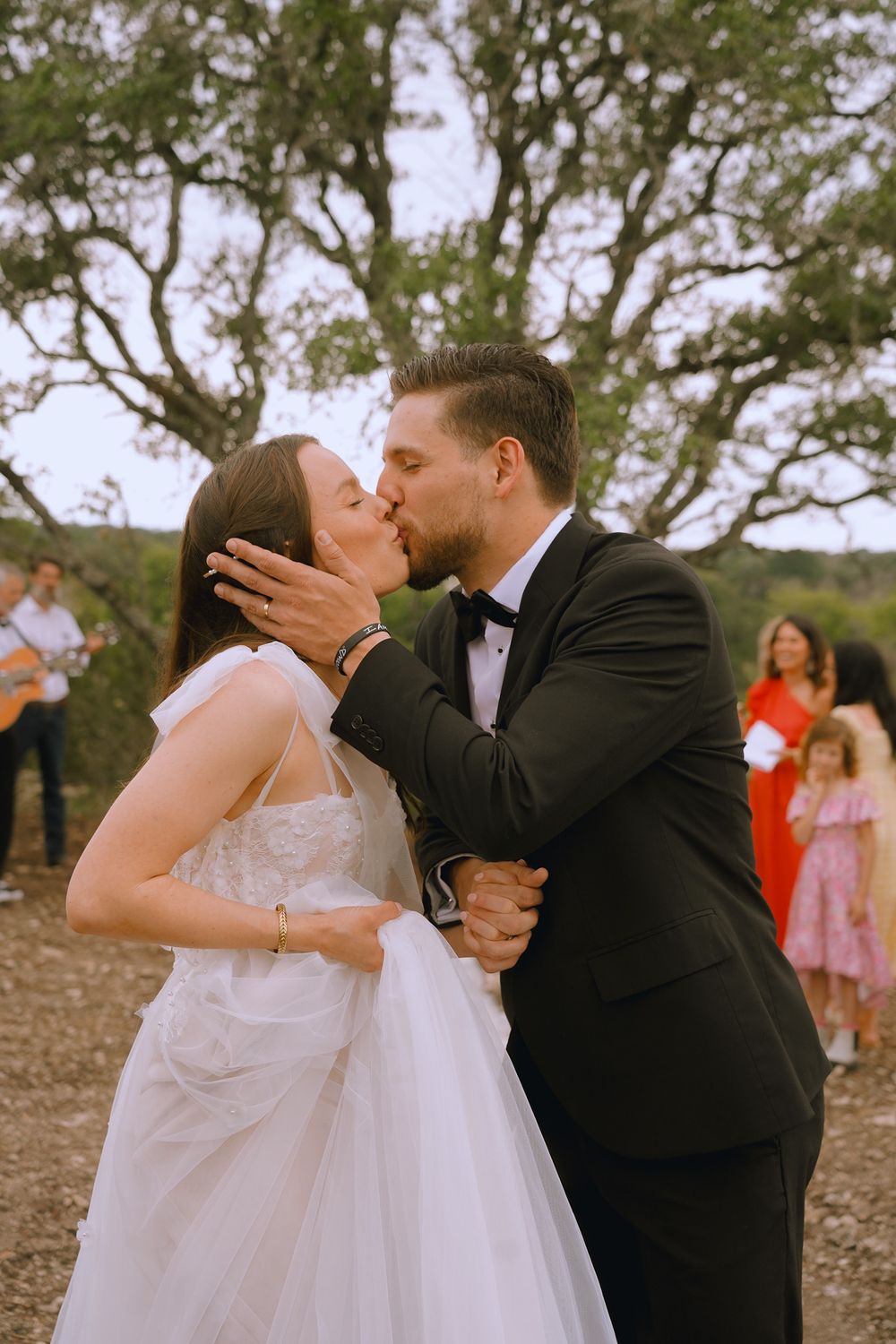 A romantic wedding kiss captured outdoors with guests in the background.
