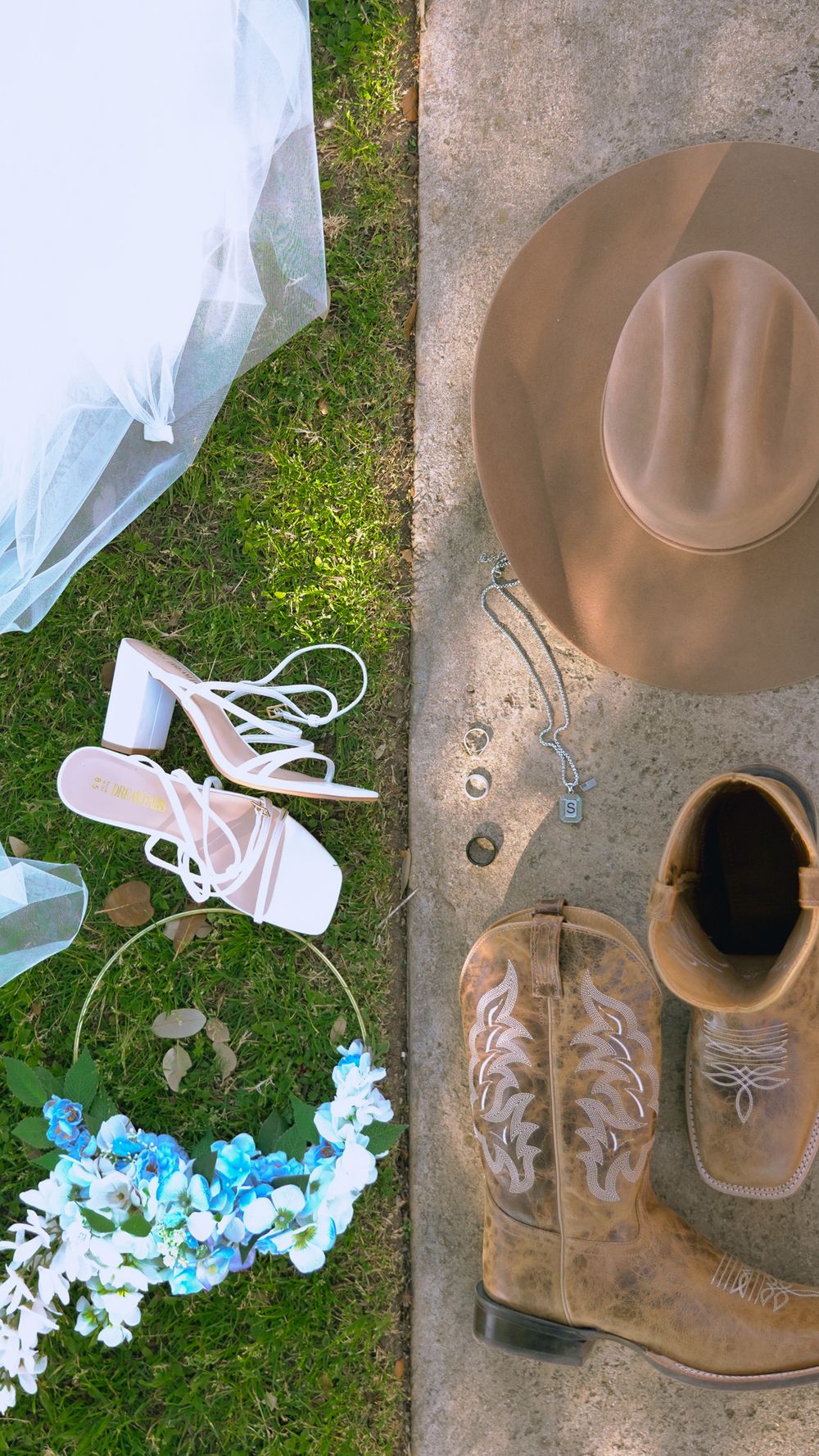 Flat lay of rustic country wedding accessories including cowboy boots, tan hat, white heels, blue floral hoop, and bridal veil.