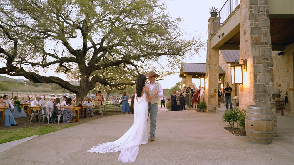 Bride and groom share first dance outside rustic Texas wedding venue with guests seated under oak tree.
