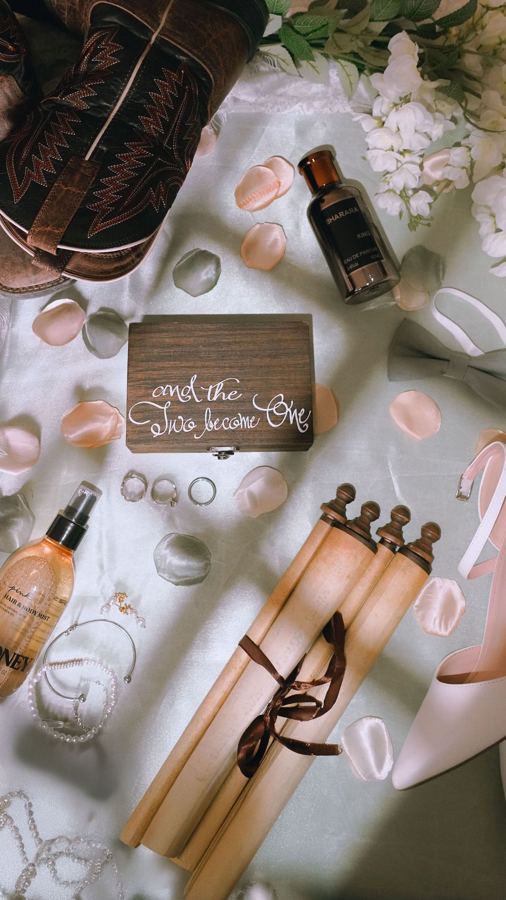 Wedding details including rings, perfume bottles, rose petals and decorative signs laid out on white fabric.
