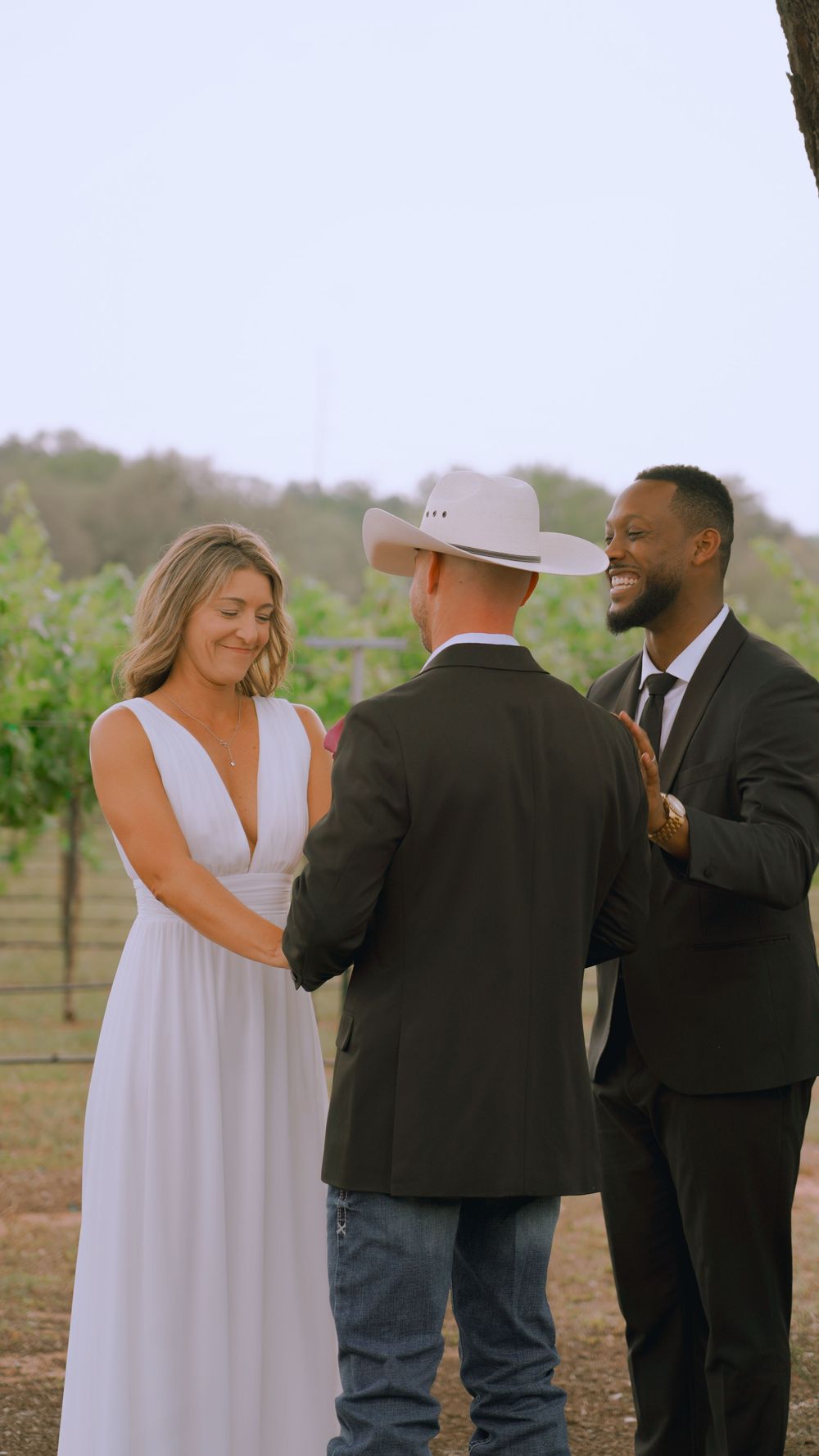 A wedding ceremony takes place in a vineyard with officiator in cowboy hat and couple exchanging vows.