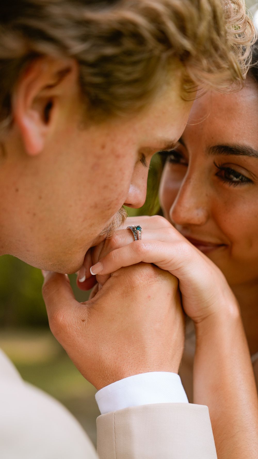 A groom in a white suit kisses his bride's hand as she smiles, wearing an engagement ring, in a romantic outdoor setting.
