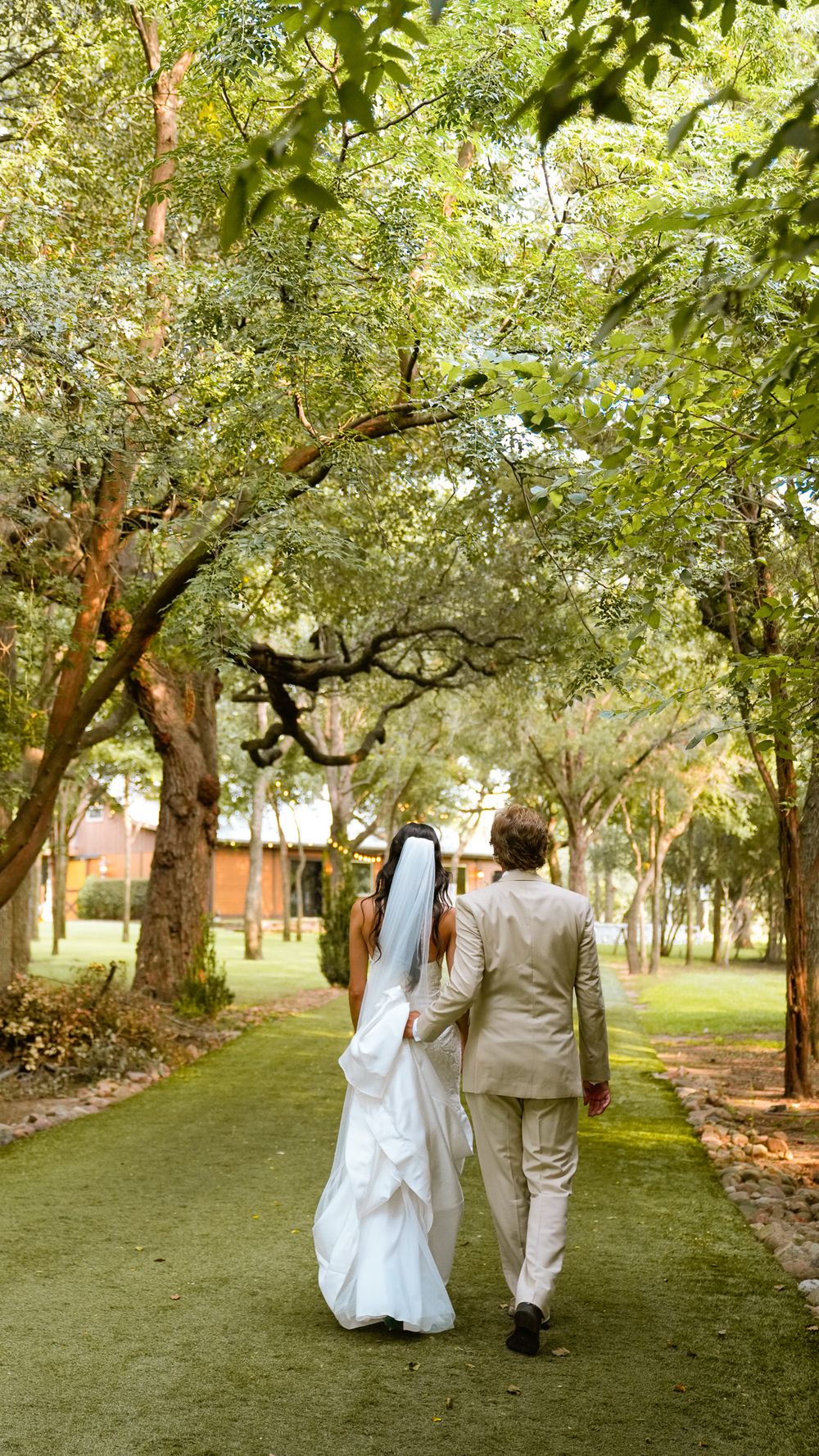 Bride and groom walking hand in hand down a lush green tree-lined path on their wedding day.
