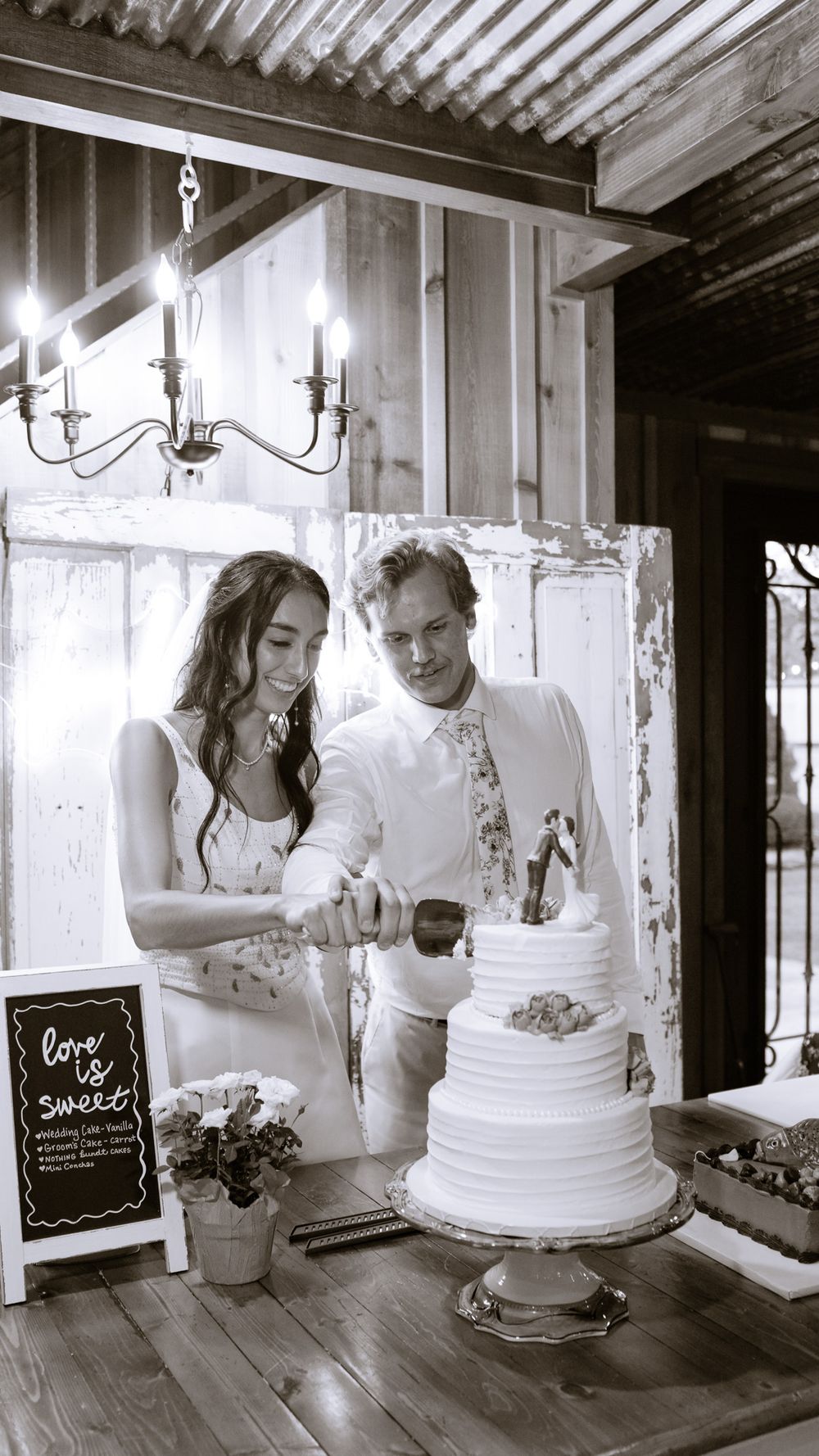 Bride and groom cut their white tiered wedding cake in black and white photo inside rustic venue.