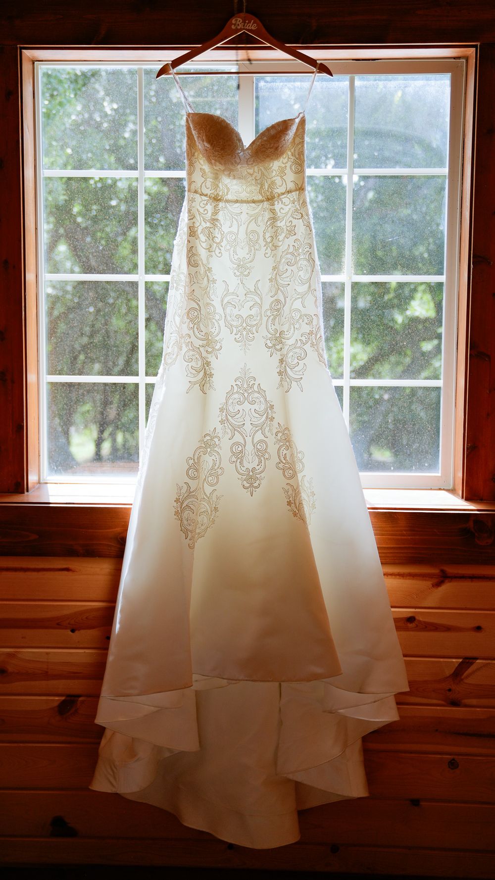 An elegant strapless white wedding gown with gold embroidery hangs in a sunlit wooden cabin window.
