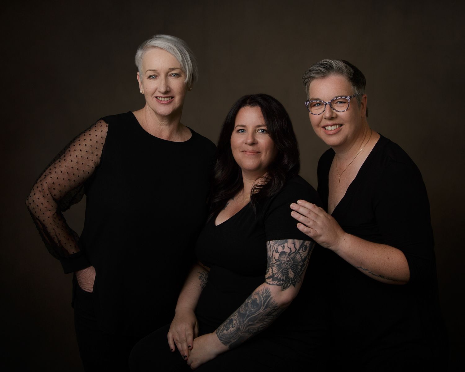Three women with tattoos standing together, smiling for a portrait, showcasing their unique body art and camaraderie.