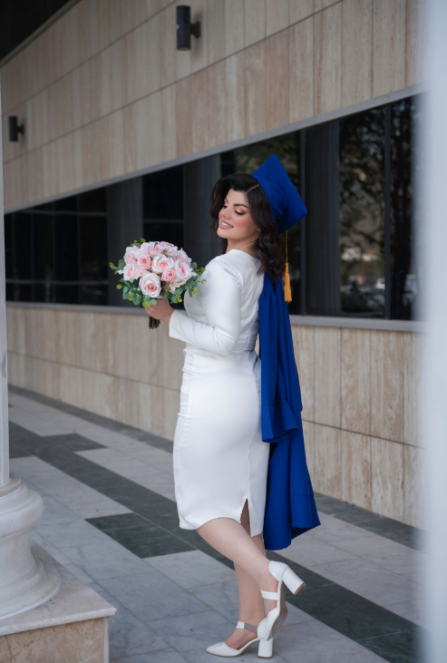 Graduate in white dress and blue cap with tassel holding pink bouquet poses by modern building exterior.