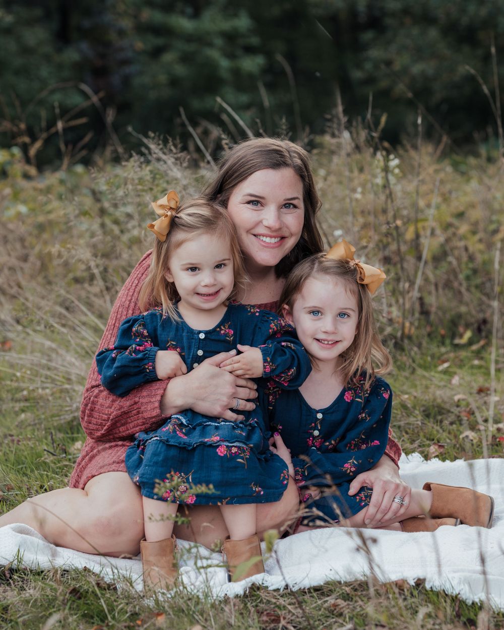 A family sits together on a blanket outdoors, wearing matching navy floral dresses and smiling warmly in a natural setting.