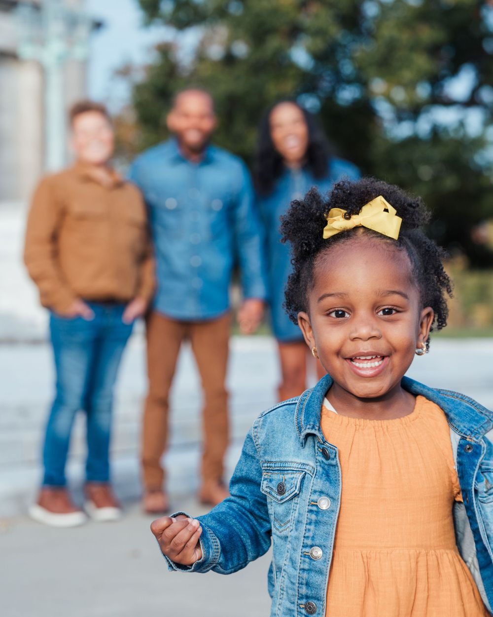 Child in denim jacket and orange dress with yellow bow smiles at camera as family members stand in background outdoors.