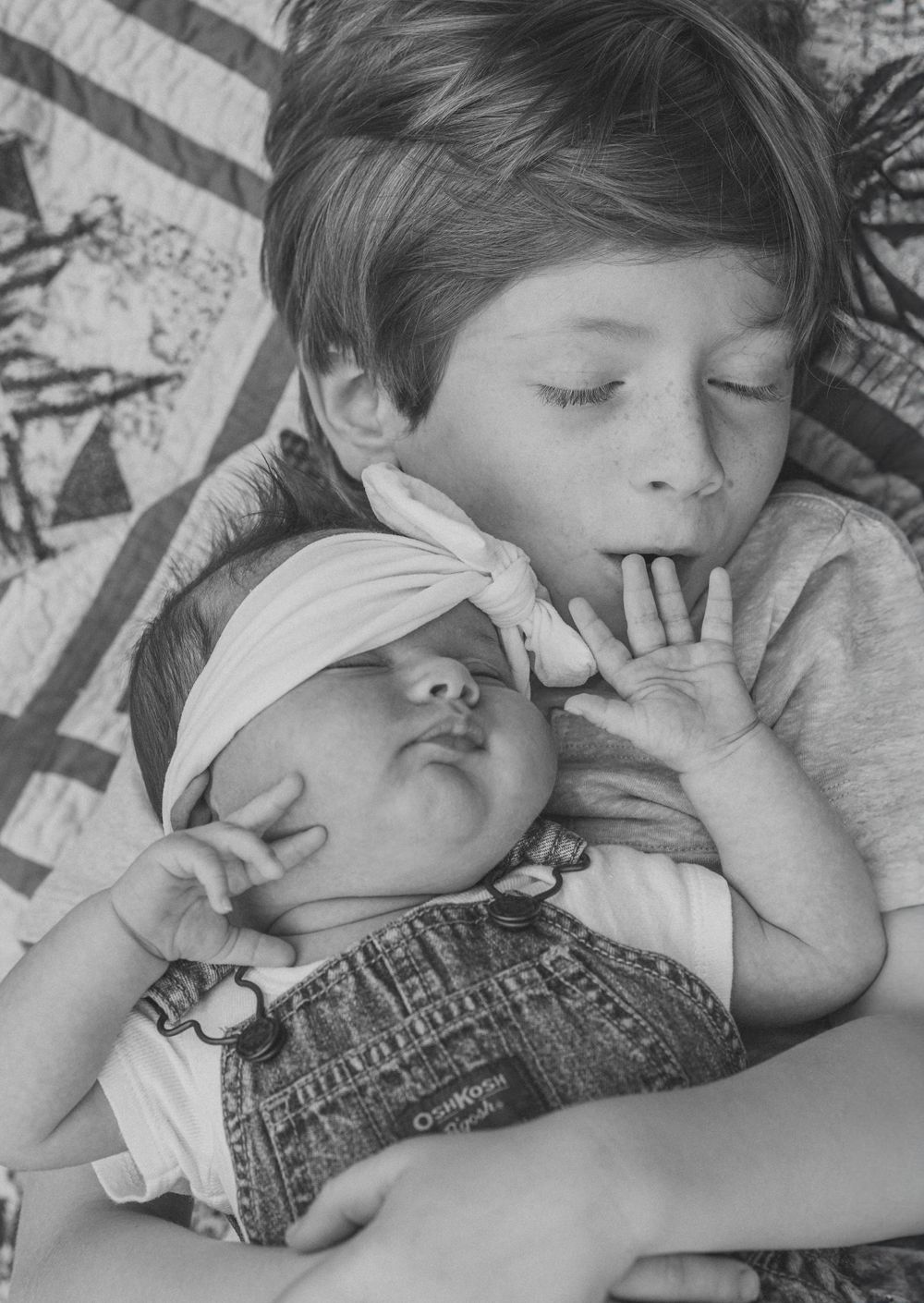 A peaceful black and white photograph shows two children sleeping together on a patterned blanket.