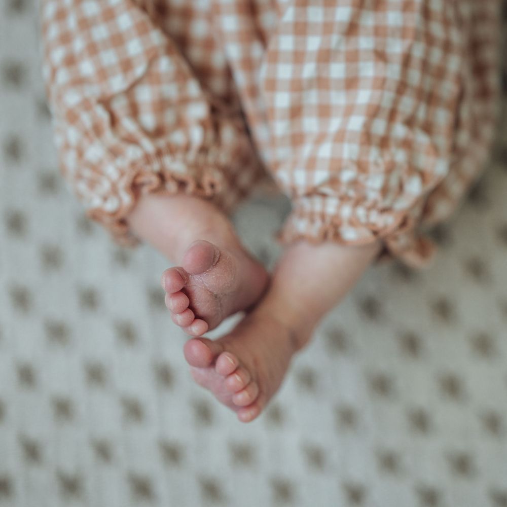 Tiny baby feet peek out from beneath orange gingham pants against a polka dot background.