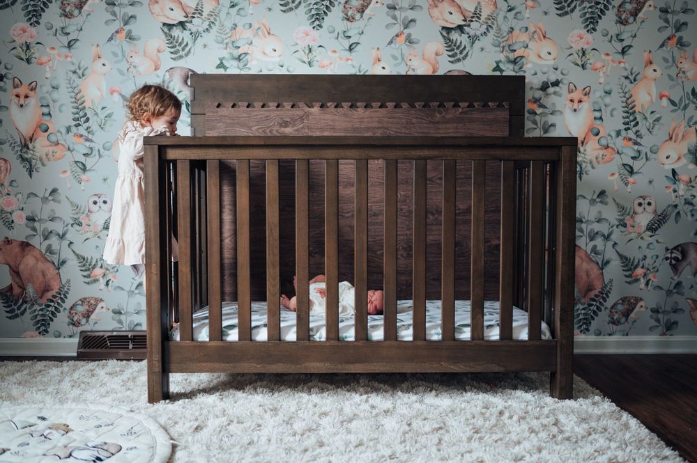 Dark wooden crib stands against floral wallpaper in nursery with white shag rug and small figure peeking from side.
