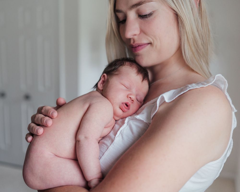 A tender moment shows a newborn baby sleeping peacefully against a soft white top in natural light.