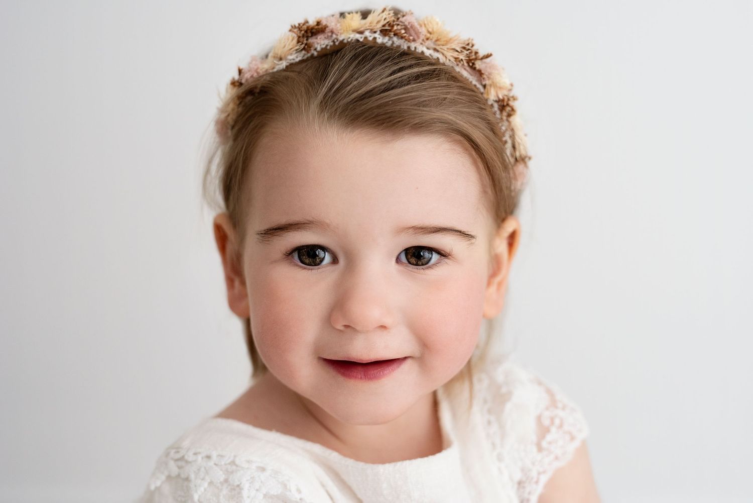 A young girl in a white lace dress and floral crown poses sweetly against a light gray backdrop.