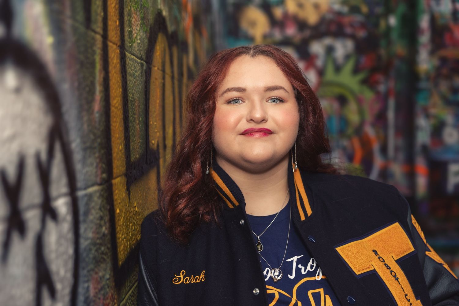A person in a varsity jacket smiles while standing against a graffiti-covered wall with soft lighting. Taken in Ann Arbor, Michigan
