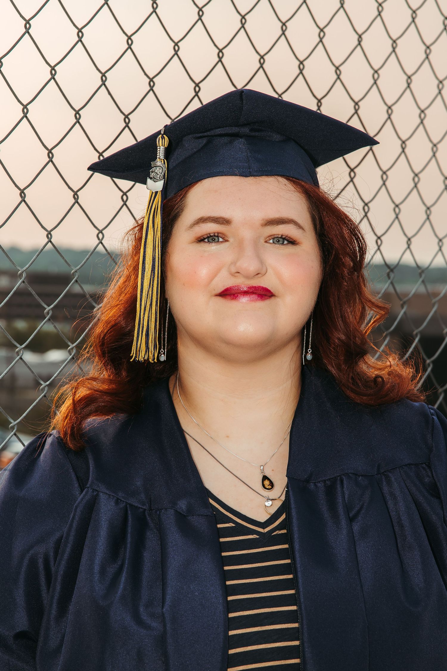 Graduate in navy cap and gown with gold tassel poses against chain link fence background.