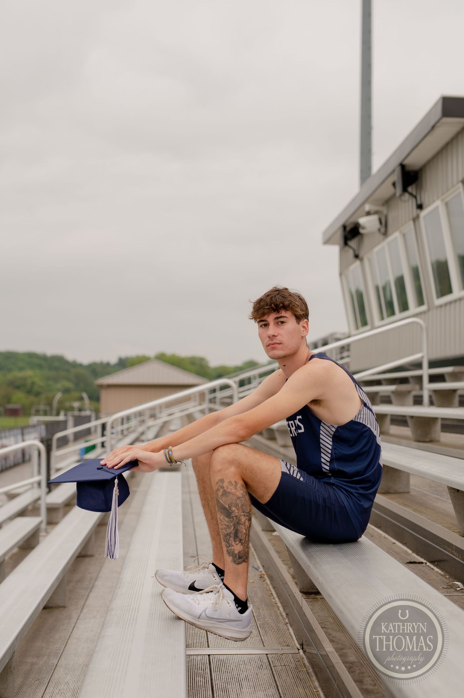 A graduate in athletic wear sits on metal bleachers holding their graduation cap.