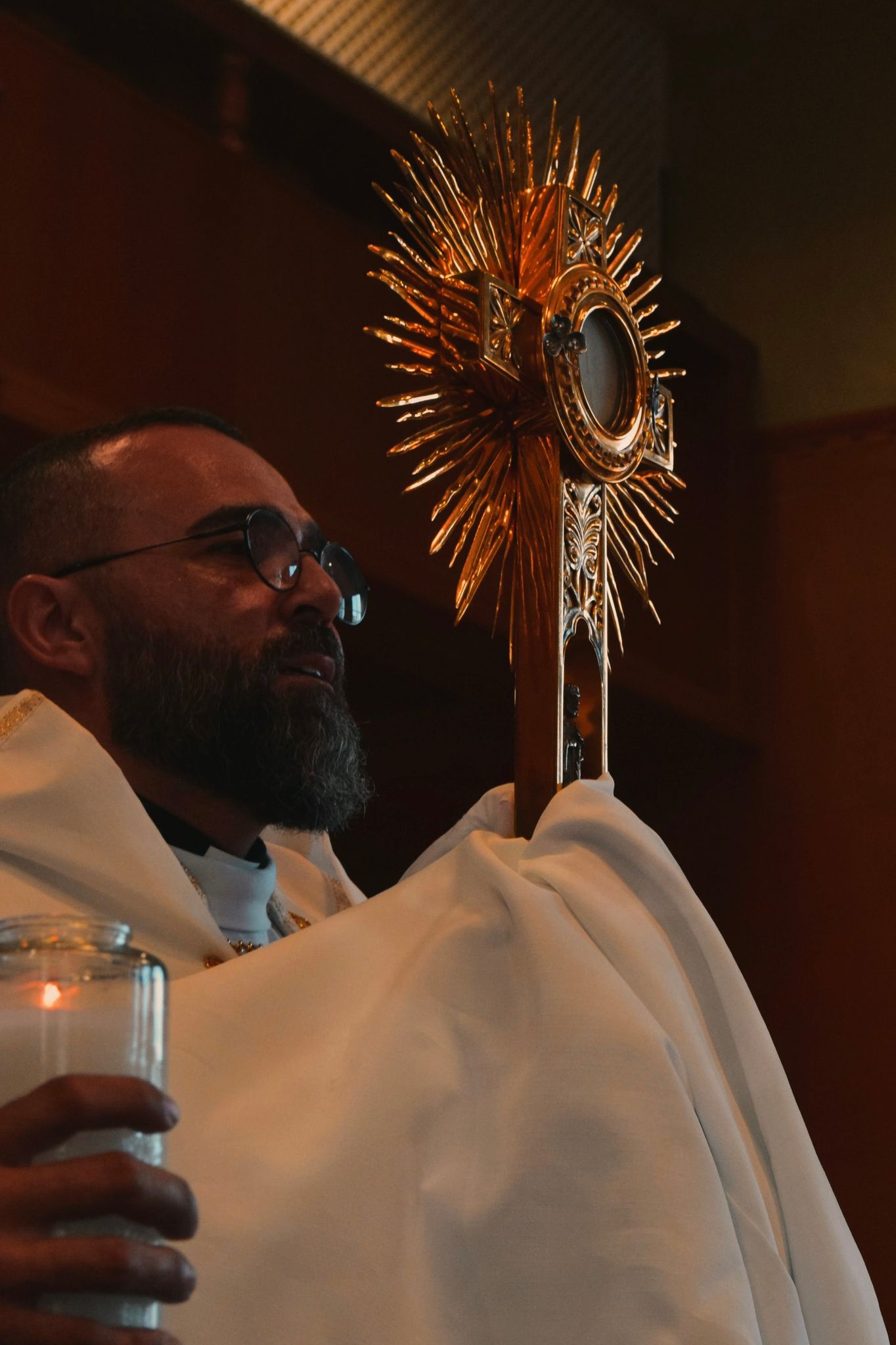 Clergyman in white robes holding ornate golden sunburst monstrance during Catholic Eucharistic adoration ceremony.