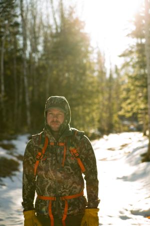 Hunter wearing camouflage gear and orange safety straps stands in sunlit snowy forest.