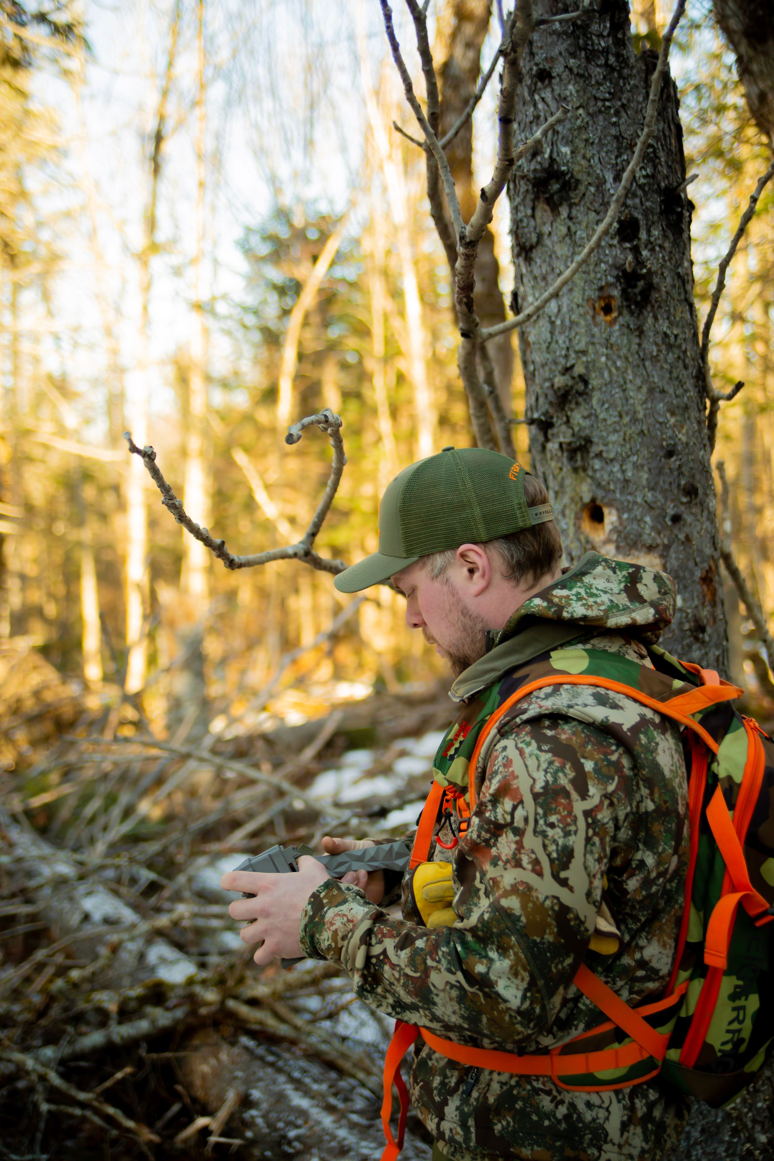 Side view of hunter in camouflage gear and green cap examining equipment in golden forest light.