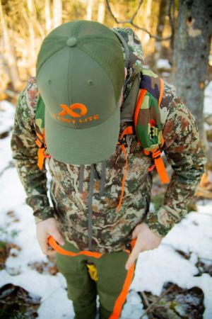 A green baseball cap with orange accents and backpack straps on camouflage hunting gear in snowy woods.