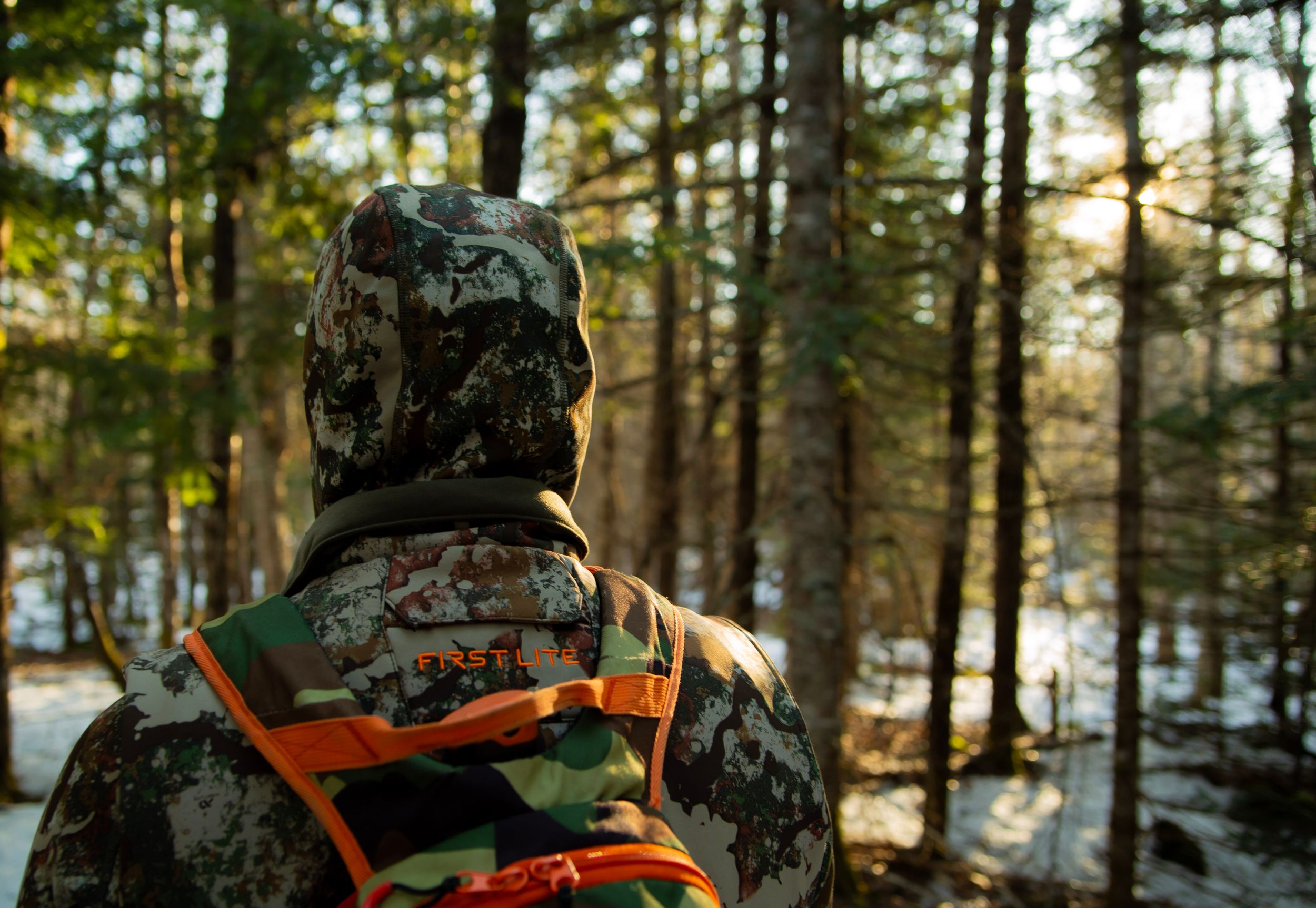 Hunter in camouflage gear with orange backpack looking out at snowy winter forest.