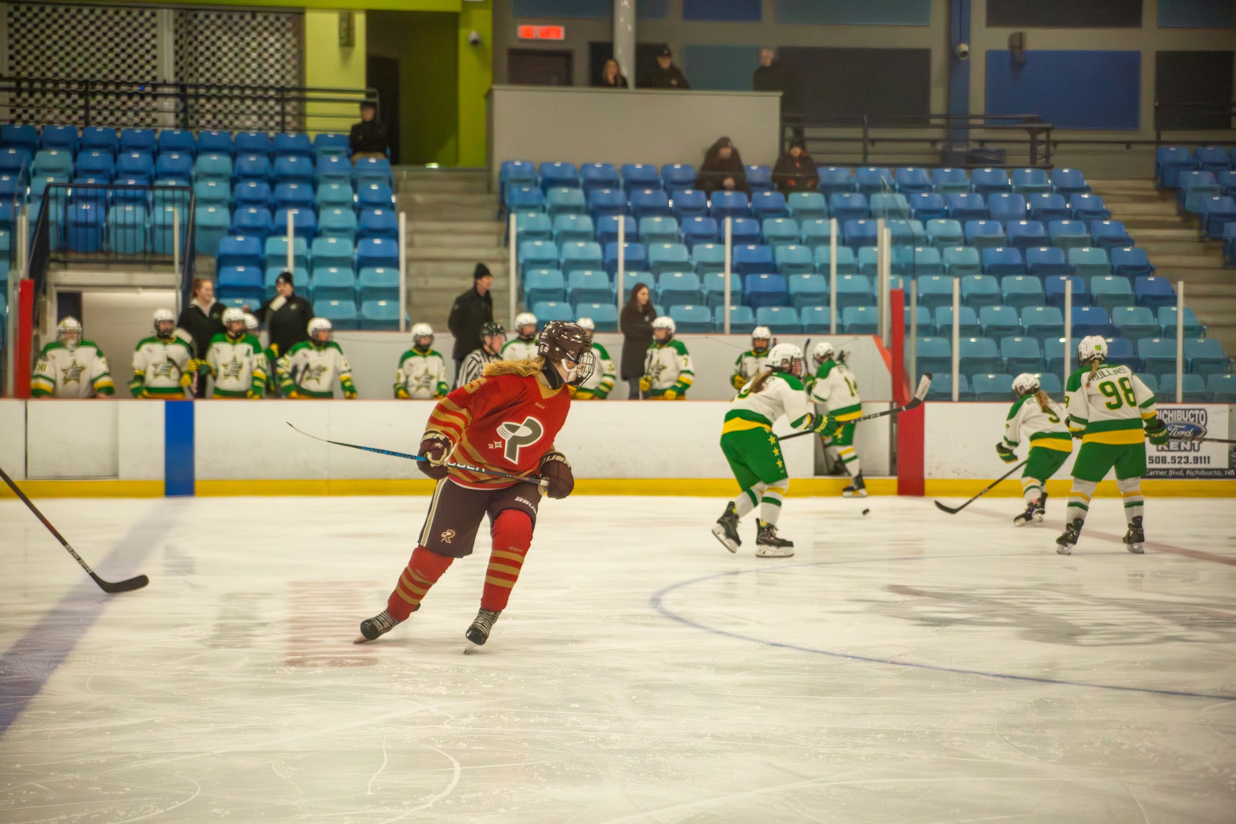 A hockey player in red jersey skates with the puck while green-uniformed opponents defend.