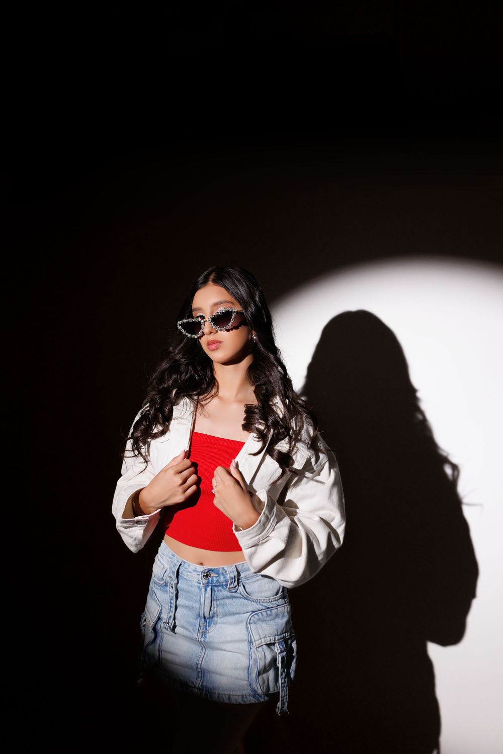 Artistic portrait against dark background with dramatic spotlight showing white jacket, red top and denim skirt outfit.