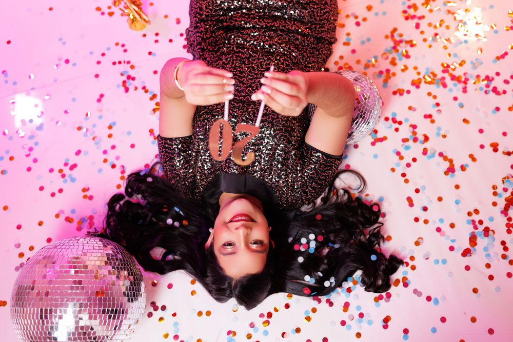 Person in sequin dress poses with confetti and disco ball on pink background for festive shoot.