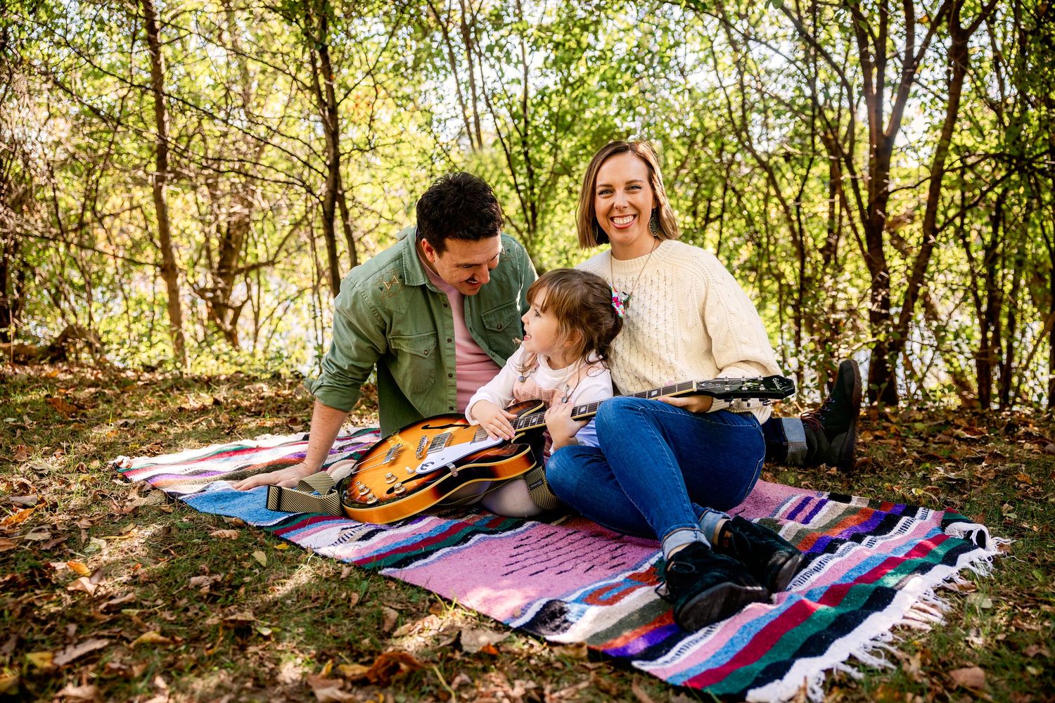 A family enjoys a peaceful afternoon on a blanket in a sunlit wooded area with a guitar at a Novi Park by Rachel Martinez Photography