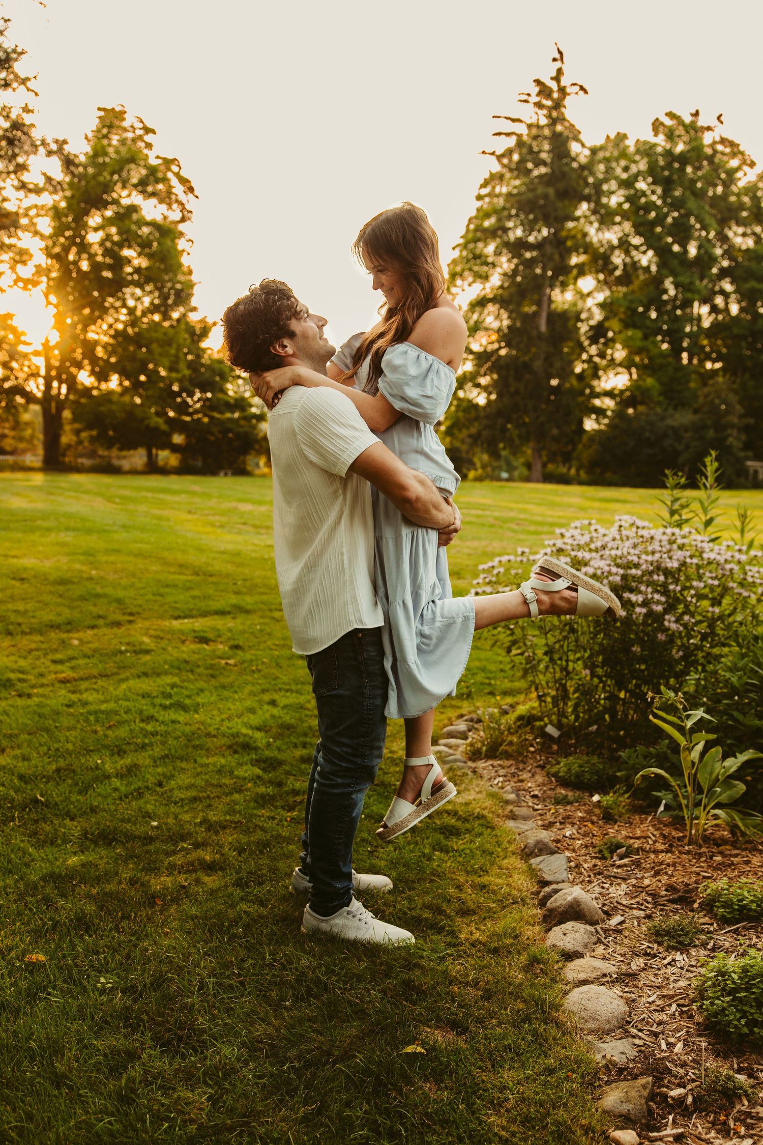 A romantic couple embraces in a garden at sunset with golden light filtering through trees in the background.